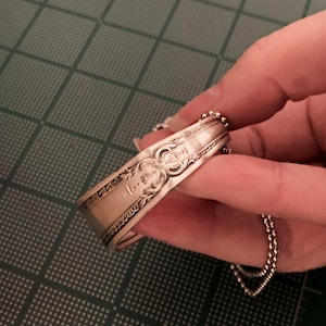 May include: A silver spoon bracelet with a decorative design and a chain attached.