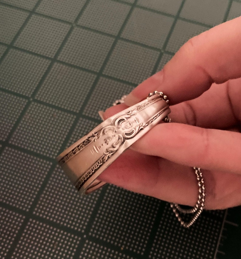 May include: A silver spoon bracelet with a decorative design and a chain attached.