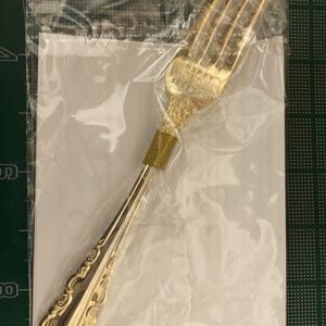 May include: A gold-colored fork with ornate detailing, sealed in a clear plastic bag. The fork's handle and head feature decorative patterns. The text "HAPPY GOLDEN" is visible on the fork's head. The fork is likely part of a flatware set.