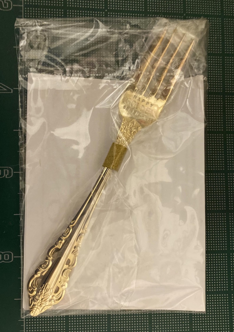 May include: A gold-colored fork with ornate detailing, sealed in a clear plastic bag. The fork's handle and head feature decorative patterns. The text "HAPPY GOLDEN" is visible on the fork's head. The fork is likely part of a flatware set.