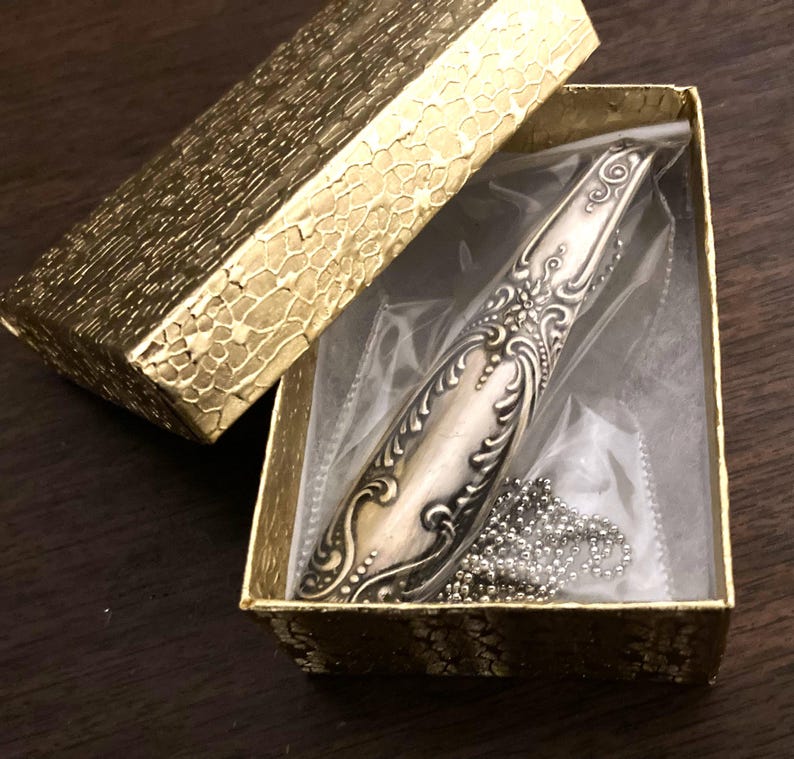 May include: A silver ornate spoon necklace in a gold box with a clear plastic insert. The spoon has a decorative design and is attached to a silver chain.