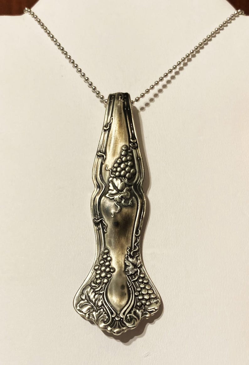 May include: A silver necklace with a pendant made from a vintage spoon. The spoon is decorated with a grapevine design.