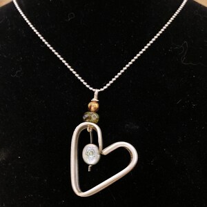 May include: A silver necklace with a heart-shaped pendant. The pendant has a small, round, iridescent bead hanging inside. The necklace also has three small, round beads in shades of brown and green.