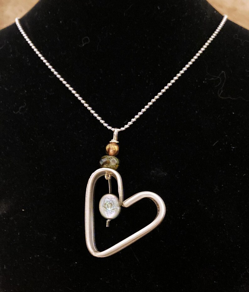 May include: A silver necklace with a heart-shaped pendant. The pendant has a small, round, iridescent bead hanging inside. The necklace also has three small, round beads in shades of brown and green.