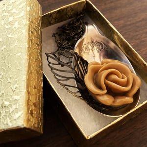 May include: A brown sugar rose charm necklace in a gold gift box. The rose is on a vintage silver spoon with a decorative design. The necklace is on a bronze chain.