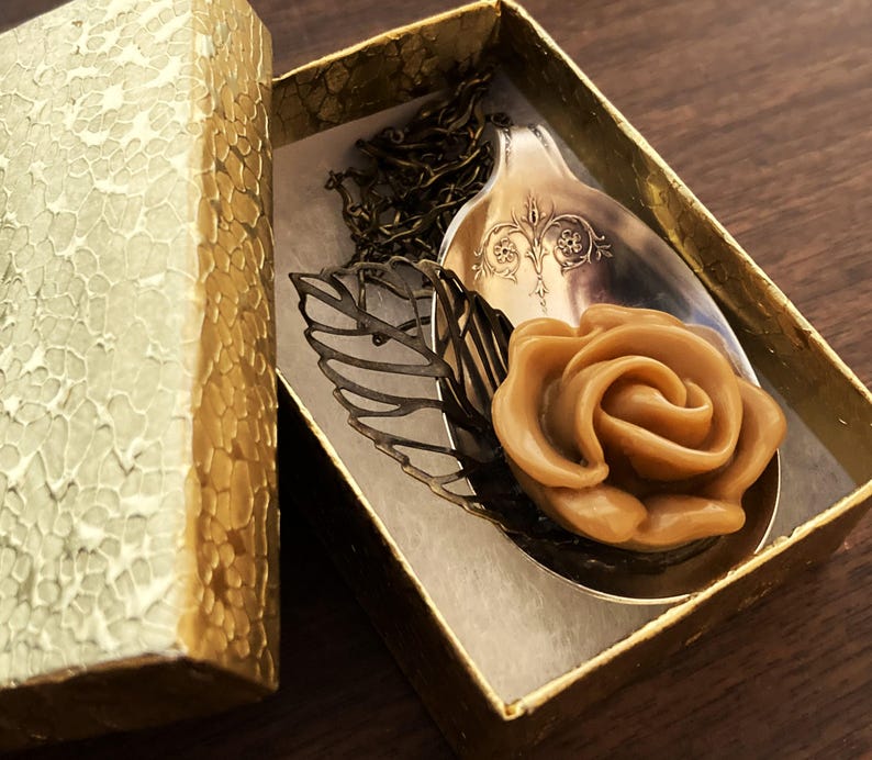 May include: A brown sugar rose charm necklace in a gold gift box. The rose is on a vintage silver spoon with a decorative design. The necklace is on a bronze chain.