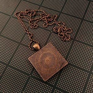 K&ouml;nnte beinhalten: Eine quadratische Anh&auml;nger-Halskette mit einem detaillierten, komplizierten Design. Der Anh&auml;nger ist aus kupferfarbenem Metall gefertigt und h&auml;ngt an einer Kette.