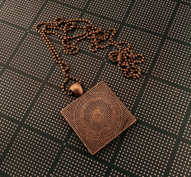K&ouml;nnte beinhalten: Eine quadratische Anh&auml;nger-Halskette mit einem detaillierten, komplizierten Design. Der Anh&auml;nger ist aus kupferfarbenem Metall gefertigt und h&auml;ngt an einer Kette.