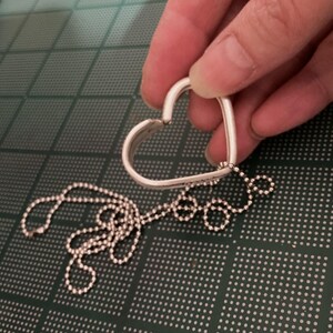 Puede incluir: Un colgante de plata en forma de coraz&oacute;n con una cadena de bolas de plata. El colgante est&aacute; abierto en la parte superior y la cadena est&aacute; unida a la parte inferior.