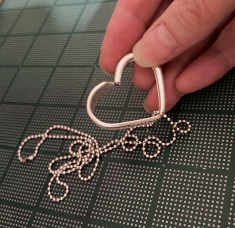 Puede incluir: Un colgante de plata en forma de coraz&oacute;n con una cadena de bolas de plata. El colgante est&aacute; abierto en la parte superior y la cadena est&aacute; unida a la parte inferior.