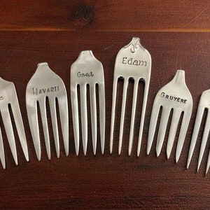 May include: Six silver forks with different cheese names engraved on them: Brie, Havarti, Goat, Edam, Gruyere, and Bleu.