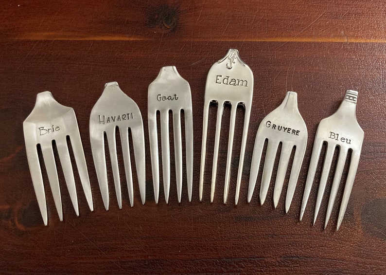 May include: Six silver forks with different cheese names engraved on them: Brie, Havarti, Goat, Edam, Gruyere, and Bleu.