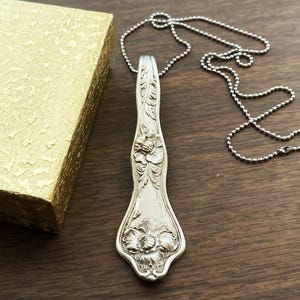 May include: A silver necklace with a pendant made from a vintage spoon. The spoon is decorated with floral designs.