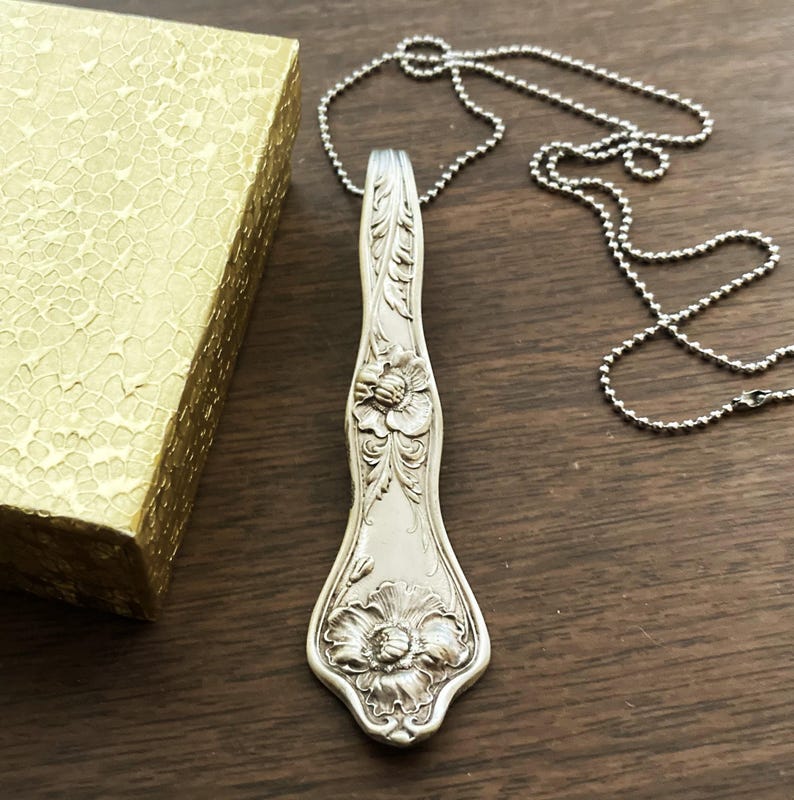 May include: A silver necklace with a pendant made from a vintage spoon. The spoon is decorated with floral designs.