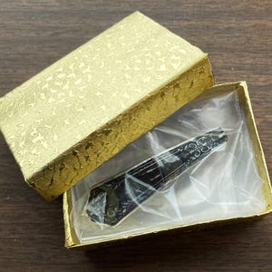 May include: A black and silver pocket knife with a decorative design on the handle, displayed in a gold gift box with a clear plastic insert.
