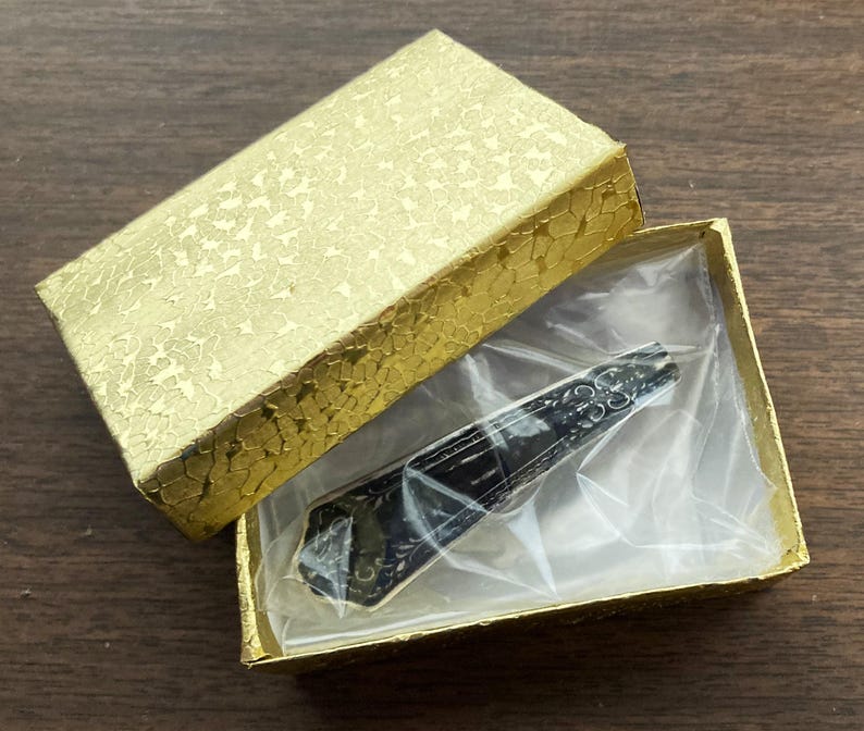 May include: A black and silver pocket knife with a decorative design on the handle, displayed in a gold gift box with a clear plastic insert.