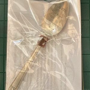May include: A silver teaspoon with the words "Coffee, Book, Repeat" engraved on the bowl. The spoon is packaged in a clear plastic bag with a white paper insert.