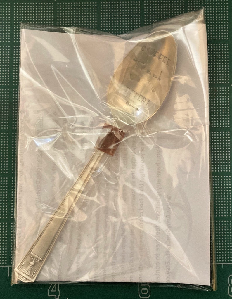 May include: A silver teaspoon with the words "Coffee, Book, Repeat" engraved on the bowl. The spoon is packaged in a clear plastic bag with a white paper insert.