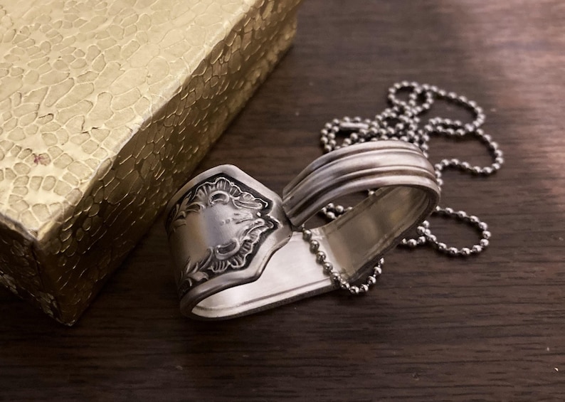 May include: A silver spoon necklace with a heart-shaped design. The spoon has a decorative pattern on the handle and is attached to a silver chain.