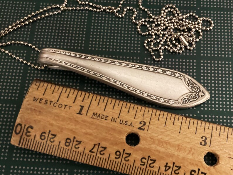 May include: A silver spoon necklace with a decorative design on the handle. The spoon is about 2 inches long and is attached to a silver ball chain.