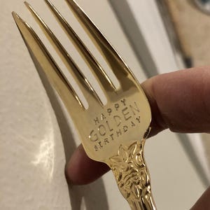May include: A gold-colored fork with the words "HAPPY GOLDEN BIRTHDAY" engraved on the handle. The fork has four tines and a decorative floral pattern on the handle.