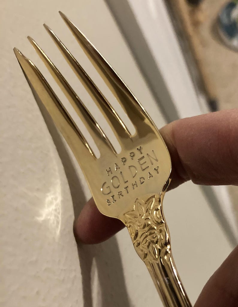 May include: A gold-colored fork with the words "HAPPY GOLDEN BIRTHDAY" engraved on the handle. The fork has four tines and a decorative floral pattern on the handle.