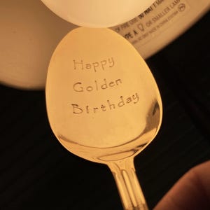 May include: A golden spoon with the words "Happy Golden Birthday" engraved on the bowl. The spoon is made of a shiny, reflective metal and is set against a dark background.