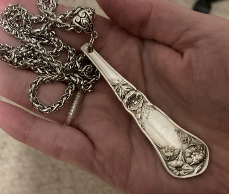 May include: A silver necklace with a long, ornate pendant made from a vintage silverware spoon. The spoon has a floral design and is attached to a silver chain.