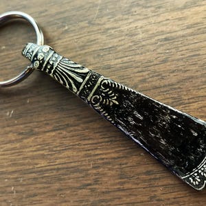 May include: A black and silver shoe horn keychain with an ornate design. The shoe horn is attached to a silver key ring.
