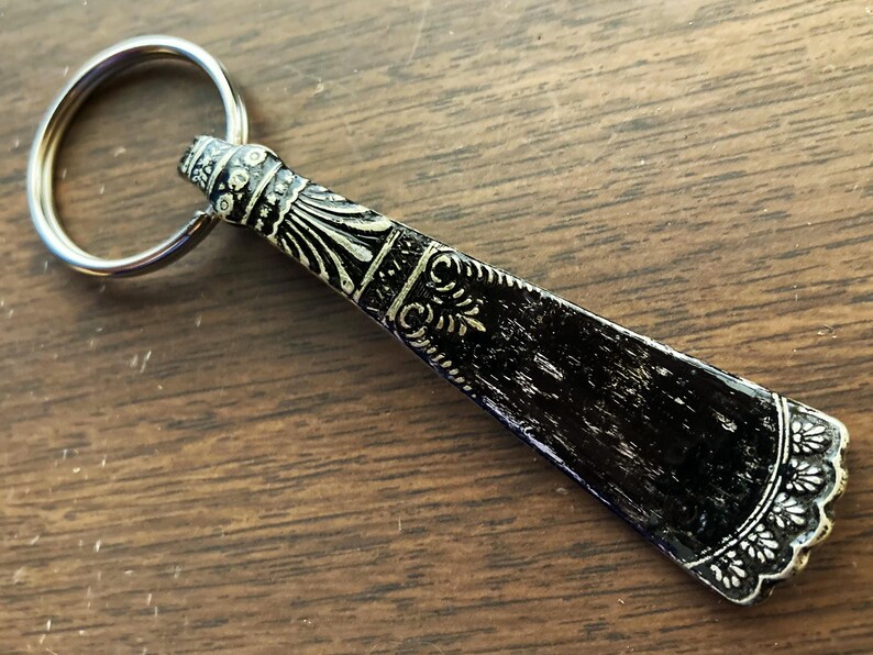 May include: A black and silver shoe horn keychain with an ornate design. The shoe horn is attached to a silver key ring.