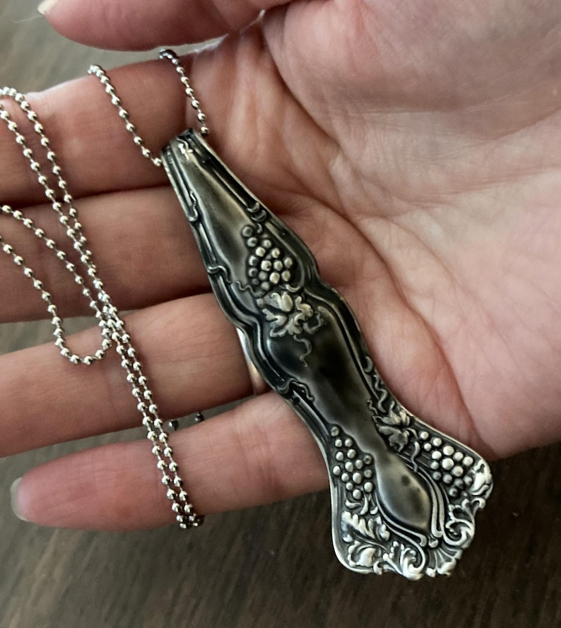 May include: A silver necklace with a vintage spoon pendant featuring a grapevine design. The spoon is oxidized and has a dark patina.