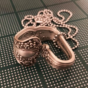 May include: A silver spoon necklace with a grapevine design. The spoon is bent into a circle and has a chain attached to it.