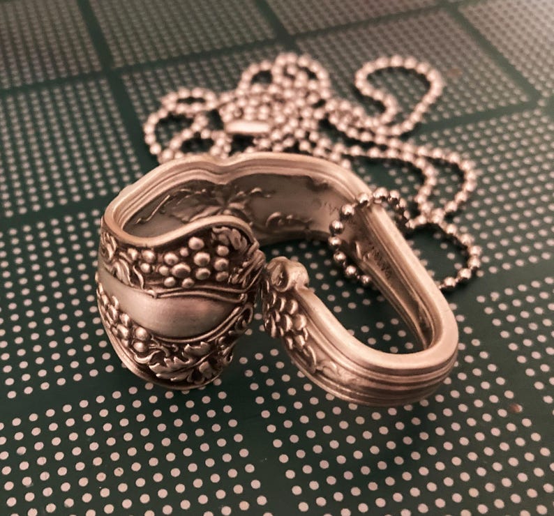 May include: A silver spoon necklace with a grapevine design. The spoon is bent into a circle and has a chain attached to it.