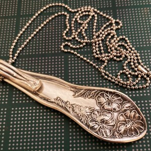 May include: A silver spoon necklace with a floral design. The spoon is bent into a pendant shape and hangs from a silver ball chain.