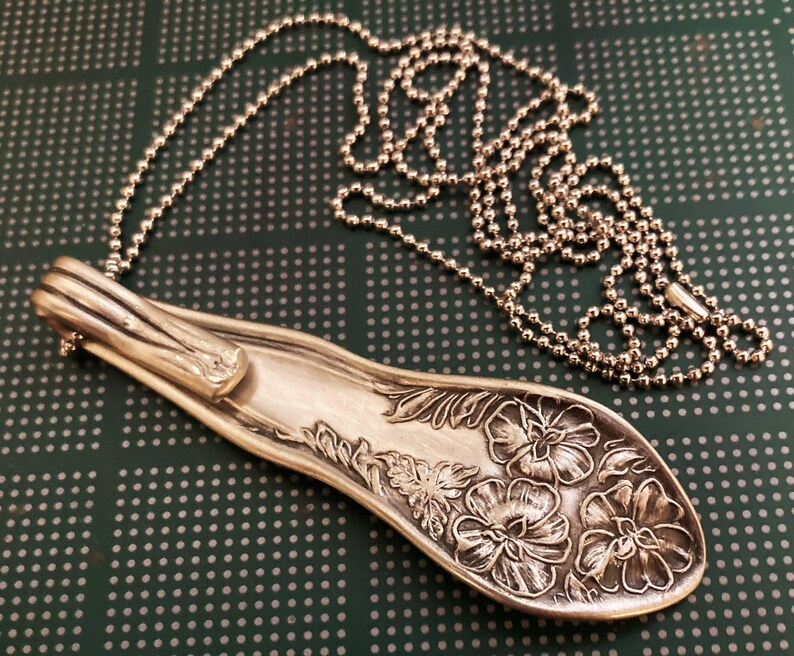 May include: A silver spoon necklace with a floral design. The spoon is bent into a pendant shape and hangs from a silver ball chain.