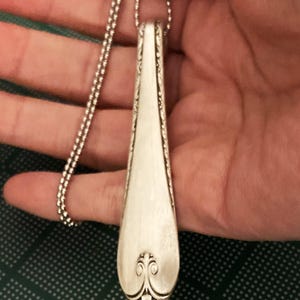 Puede incluir: Un collar con un colgante de cuchara de plata con un dise&ntilde;o floral decorativo en la parte inferior. La cuchara est&aacute; unida a una cadena de plata.