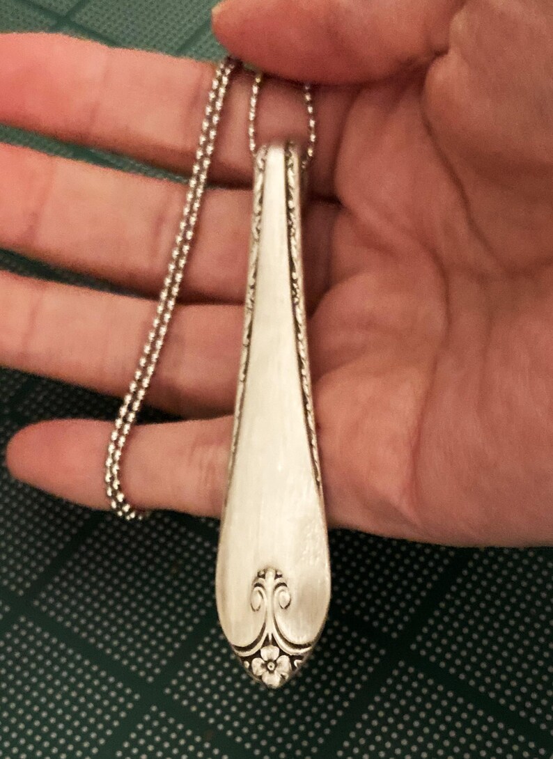 Puede incluir: Un collar con un colgante de cuchara de plata con un dise&ntilde;o floral decorativo en la parte inferior. La cuchara est&aacute; unida a una cadena de plata.
