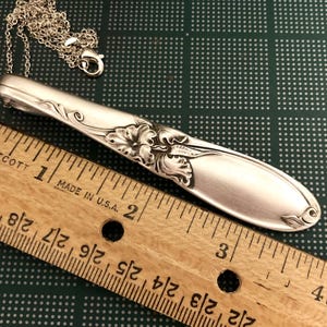 Puede incluir: Colgante de collar plateado hecho de una cuchara, con detalles florales y de hojas. El colgante est&aacute; unido a una delicada cadena. Una regla de madera con medidas en pulgadas est&aacute; en primer plano.