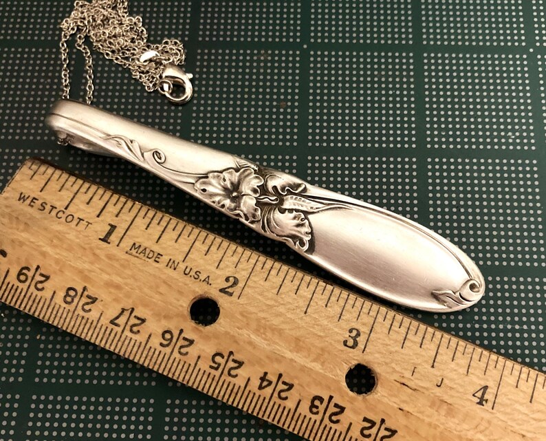 Puede incluir: Colgante de collar plateado hecho de una cuchara, con detalles florales y de hojas. El colgante est&aacute; unido a una delicada cadena. Una regla de madera con medidas en pulgadas est&aacute; en primer plano.