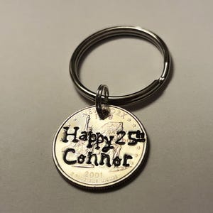 May include: A silver keychain with a round pendant. The pendant is a quarter coin with the words "Happy 25th Connor" engraved on it. The coin also features the Statue of Liberty and the year 2001.