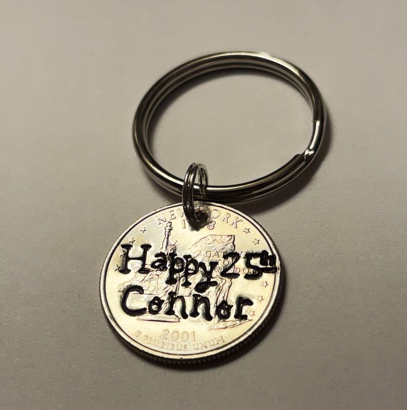 May include: A silver keychain with a round pendant. The pendant is a quarter coin with the words "Happy 25th Connor" engraved on it. The coin also features the Statue of Liberty and the year 2001.