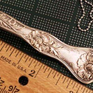 May include: A silver spoon necklace with a floral design. The spoon is about 2 inches long and has a large flower at the end. The spoon is attached to a silver chain.