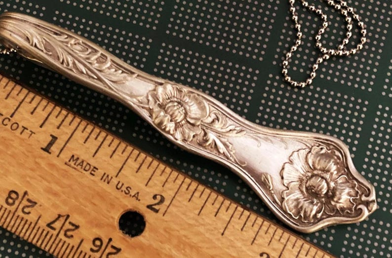 May include: A silver spoon necklace with a floral design. The spoon is about 2 inches long and has a large flower at the end. The spoon is attached to a silver chain.
