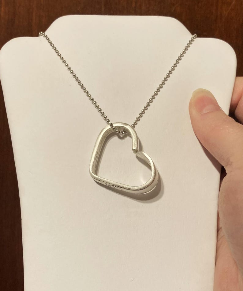 Puede incluir: Un collar de plata con un colgante en forma de coraz&oacute;n. El colgante est&aacute; hecho de una sola pieza de metal que se dobla en forma de coraz&oacute;n. La cadena es una cadena de bolas simple.