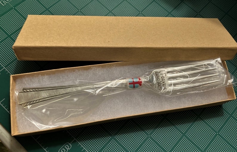 Op de afbeelding: Een zilveren vork met de woorden "Forking Love You" gegraveerd op het handvat. De vork is verpakt in een bruine kartonnen doos met wit tissuepapier.