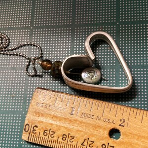 May include: A silver heart-shaped pendant necklace with a small, round, iridescent button inside. The necklace is made of a silver chain and has a brown and green bead.