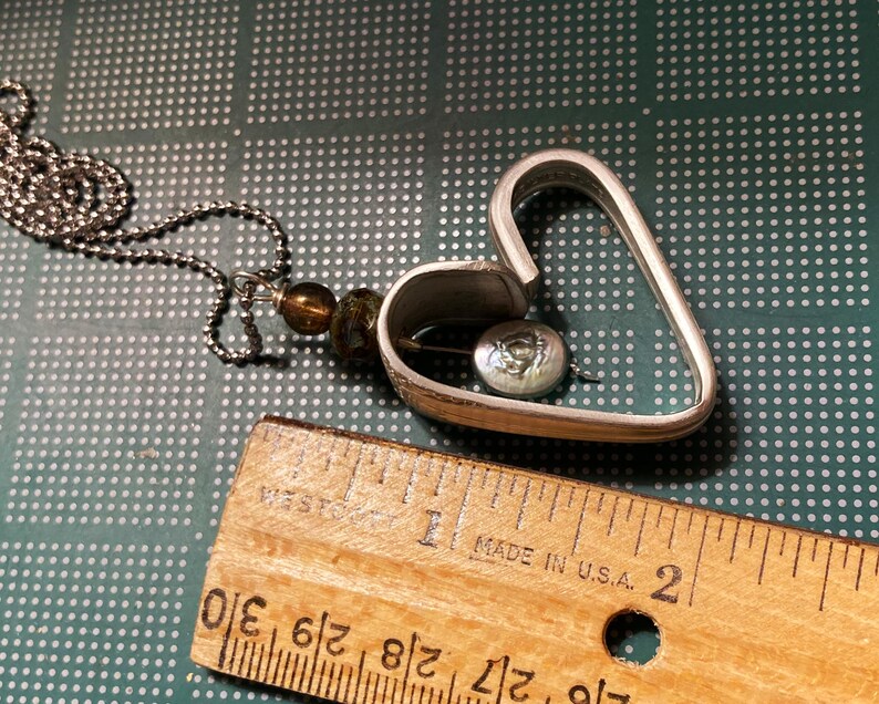 May include: A silver heart-shaped pendant necklace with a small, round, iridescent button inside. The necklace is made of a silver chain and has a brown and green bead.