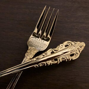 May include: Two gold-colored forks with ornate detailing, one with the words "Happy Golden Birthday" engraved on the tines. The forks are crossed against a dark wooden surface, showcasing their reflective surfaces.