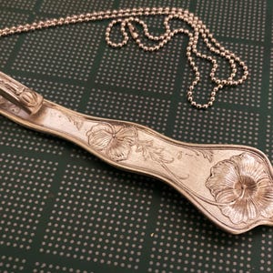 May include: A silver spoon necklace with a floral design. The spoon is bent into a loop at the top and has a chain attached to it. The spoon is decorated with a flower and leaves.