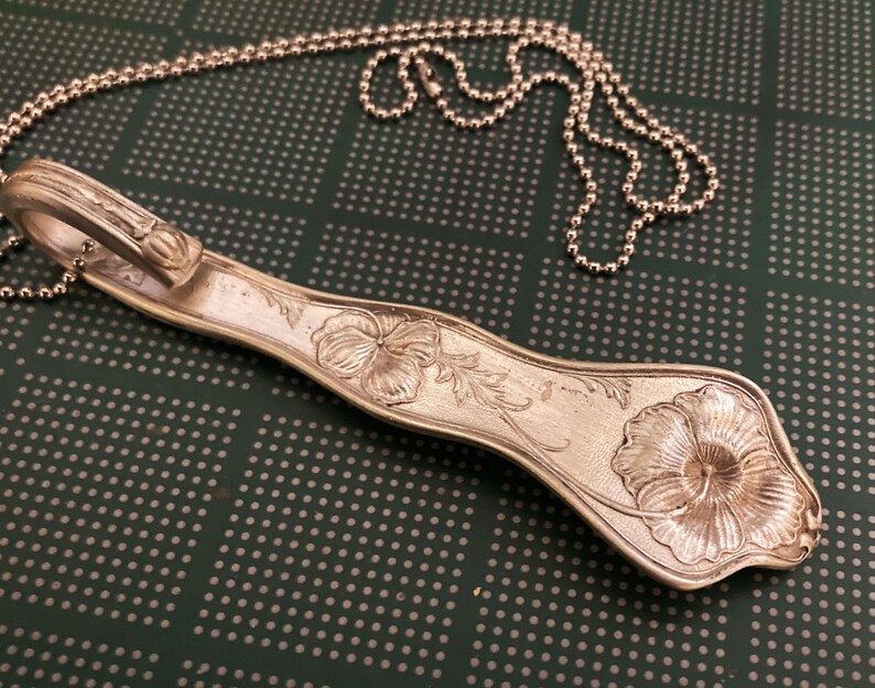 May include: A silver spoon necklace with a floral design. The spoon is bent into a loop at the top and has a chain attached to it. The spoon is decorated with a flower and leaves.